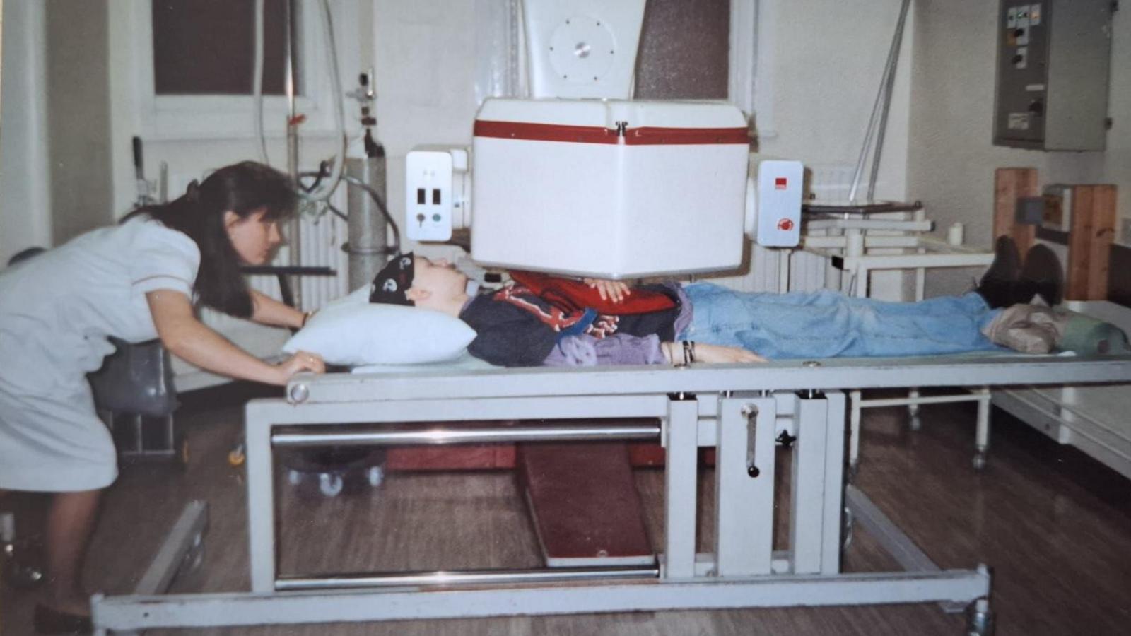 A nurse with long brown hair bends down to adjust a bed which David Wragg is lying on as he has an X-ray in a hospital during cancer treatment