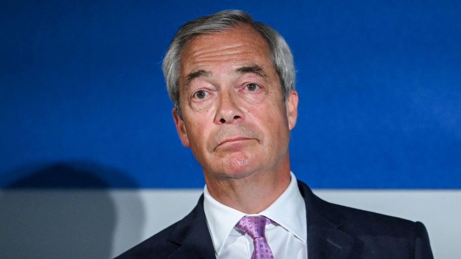 Nigel Farage looking disappointed and facing the camera. He is wearing a black blazer, white shirt and purple tie.