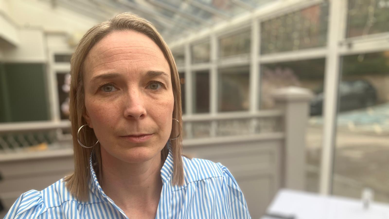 A head and shoulders picture of a woman with shoulder length blonde hair stood in a conservatory