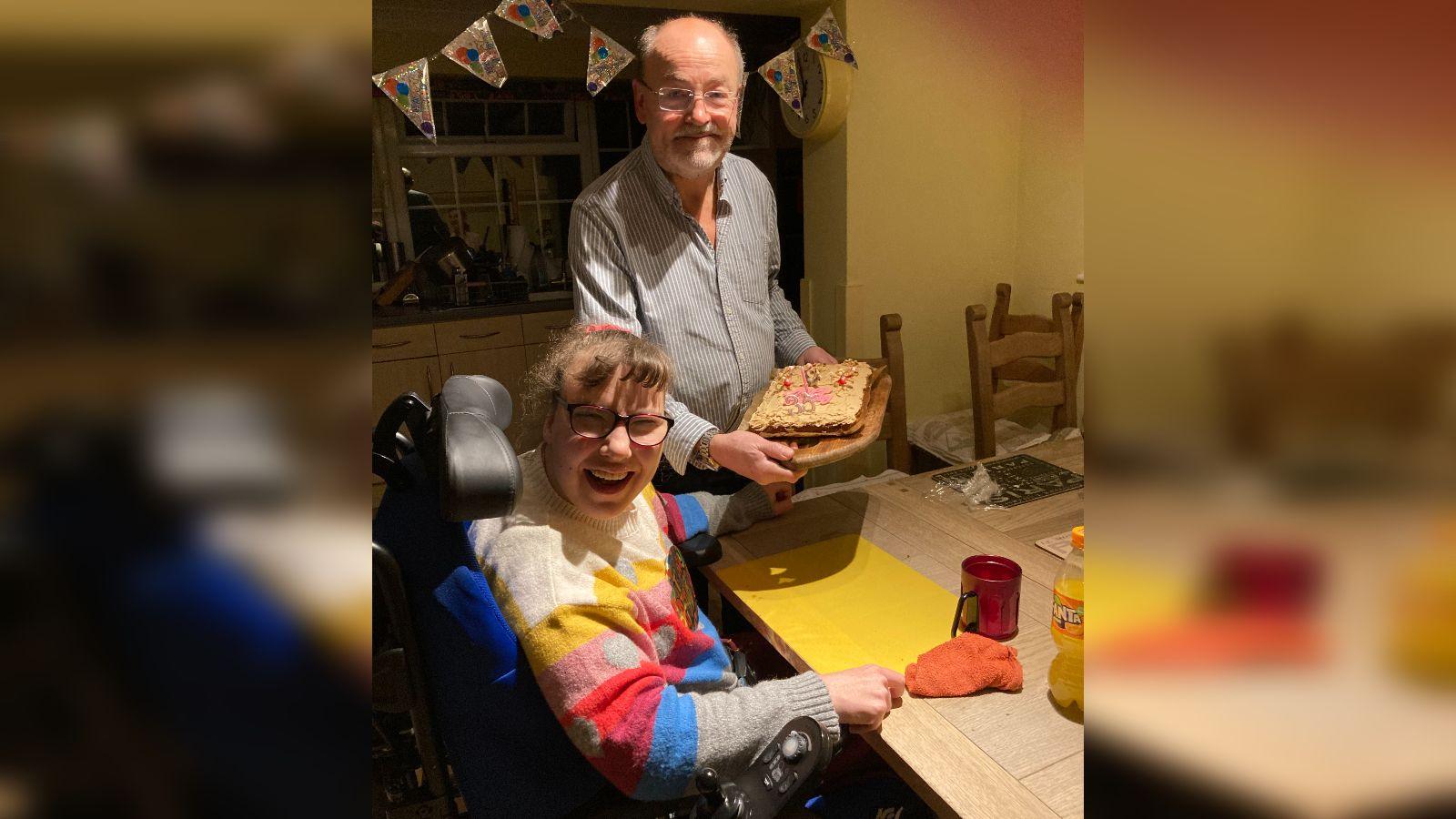 A man holds a cake smiling as a girl in a wheelchair laughs.