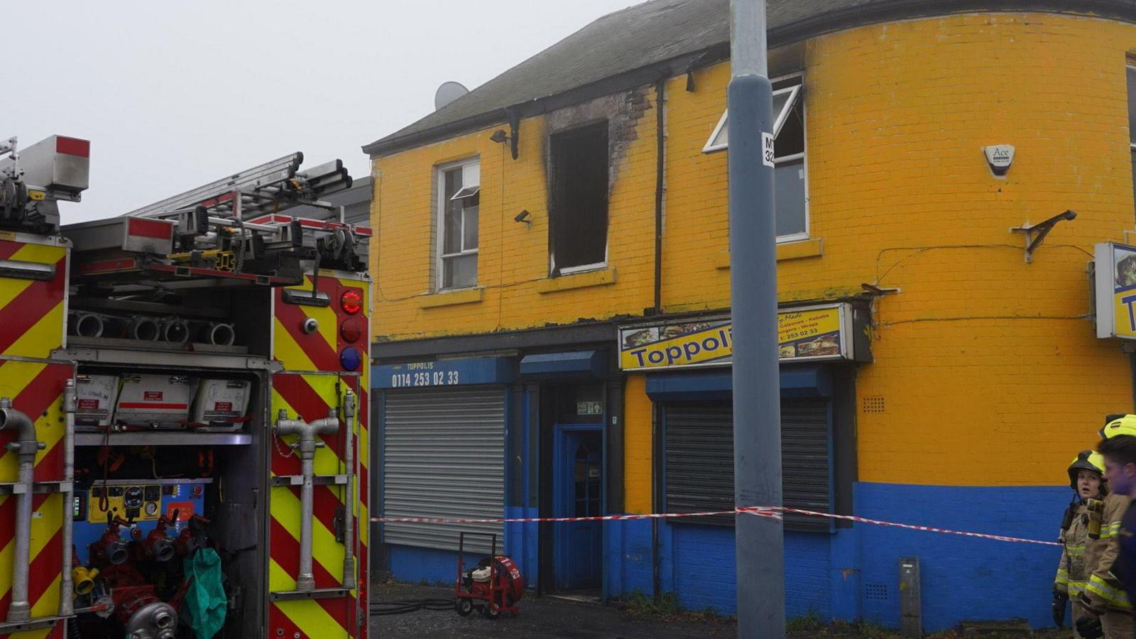 A yellow building with 'Toppolis' on a sign. There are firefighters next to the property, which has scorch marks near the windows