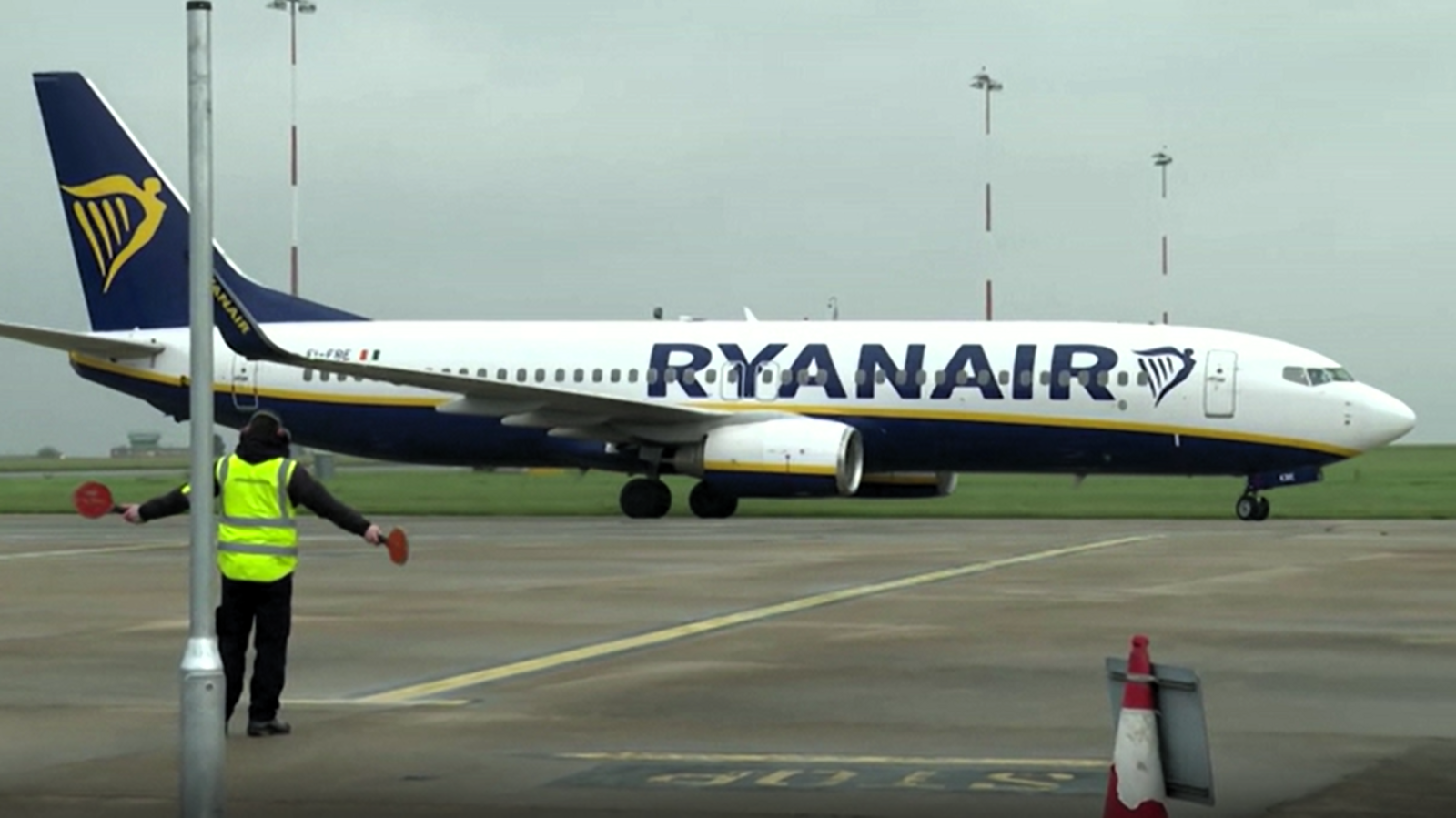 A white and blue aeroplane moves along an airport runway with a member of staff seen in the foreground in a hi-vis jacket giving instructions using red paddles.