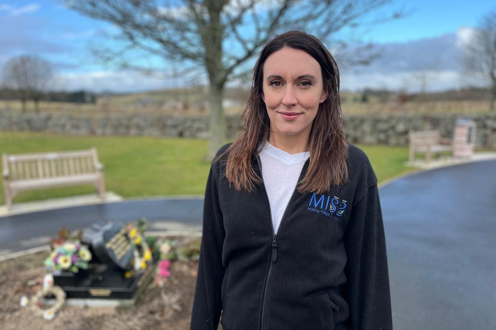 Woman, Abi Clarke, in a memorial garden, smiling and looking at camera.