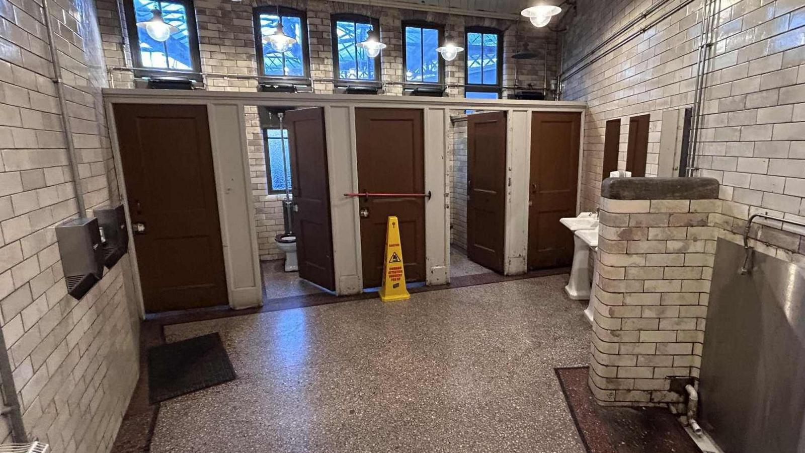 The inside of the toilets. There are five cubicles with brown doors. One of them is barricaded shut with a red pole over the front and a yellow wet floor sign in front of it. There are sinks to the right a large silver urinal in front of it. The walls are covered in greyish tiles.