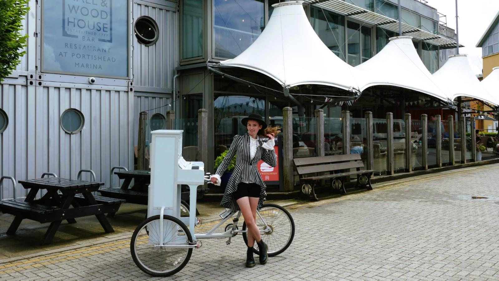 Chloe Marie Aston stands next to her piano bike outside a modern, steel-framed restaurant at a marina in Portishead, Bristol. She is wearing a black and white striped outfit with a black top hat and black boots.