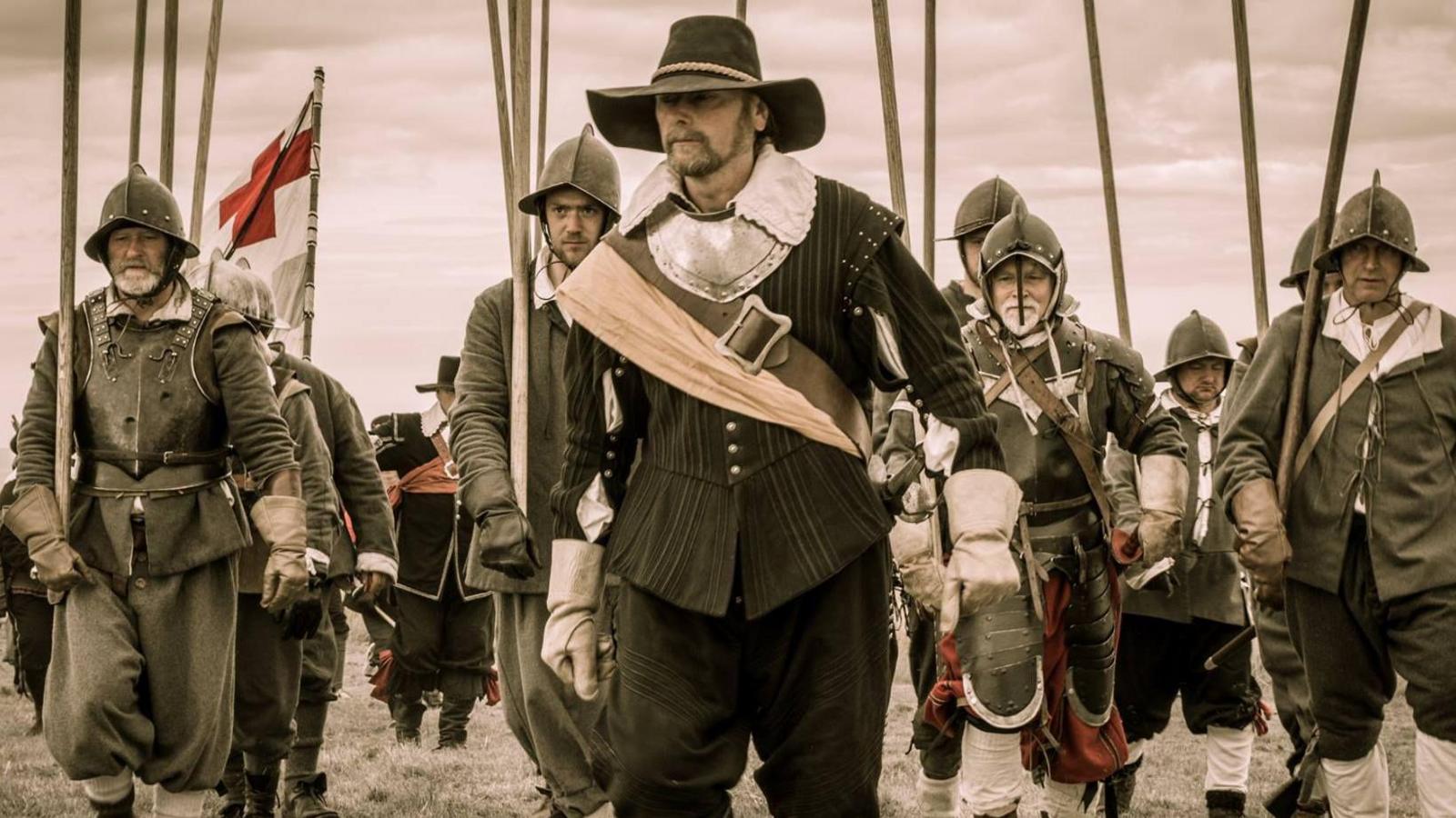An English Civil Ware enactment, showing an image of several soldiers, wearing historic outfits, armour, holding sticks. They all have hats or helmets on. 
