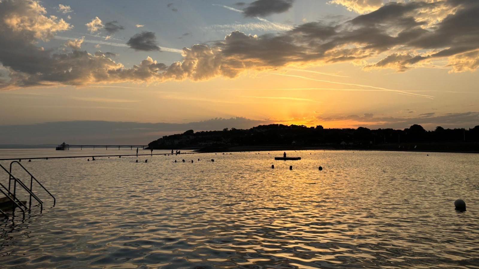 The sun is seen rising over Clevedon Marine Lake at dawn. The water is calm and the sun is lighting up the horizon beyond the pier with bright orange hues. There are some swimmers in the water, visible only as silhouettes.