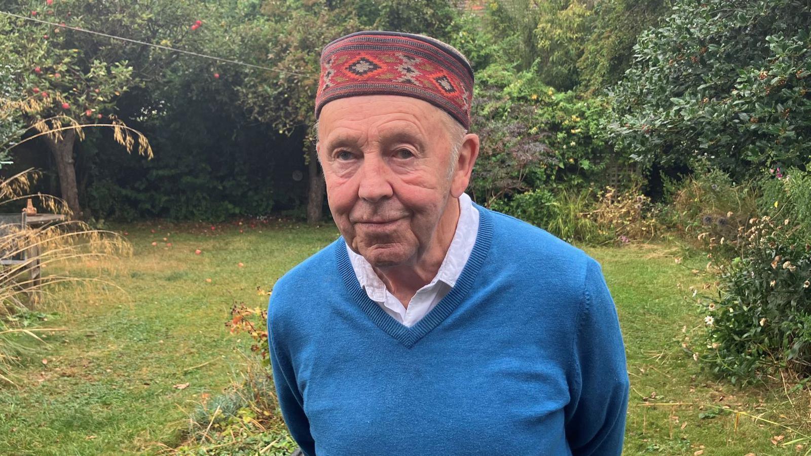 Mark Patterson, who is standing in his garden. He is wearing a blue jumper with a white shirt underneath. He also has a red patterned hat on his head.