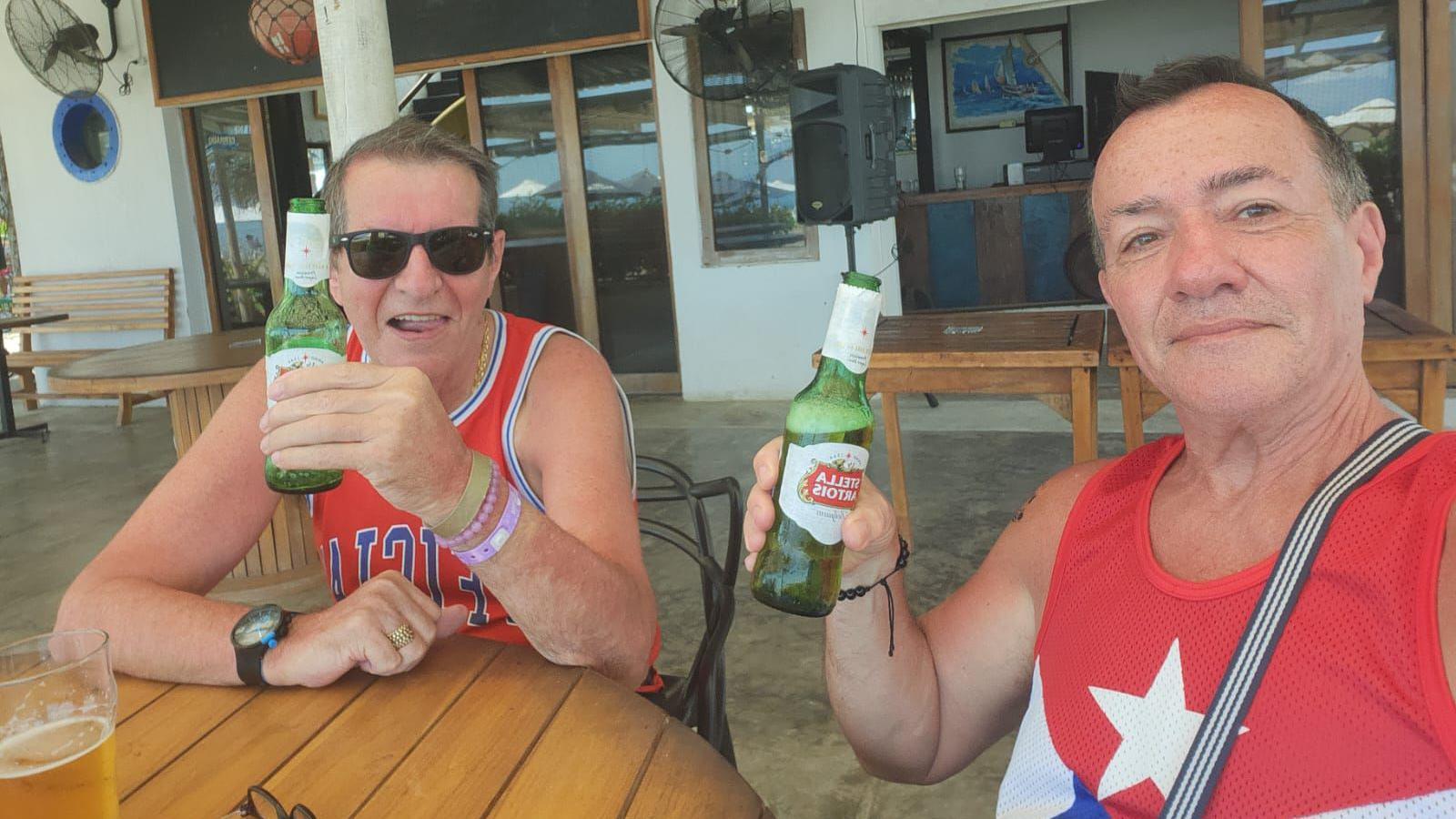 Two men sat at a table holding green beer bottles. The man on the left has short grey hair. He is wearing black sunglasses and is smiling. The man on the right has short dark hair and is smiling at the camera. Both of them are wearing red vests.