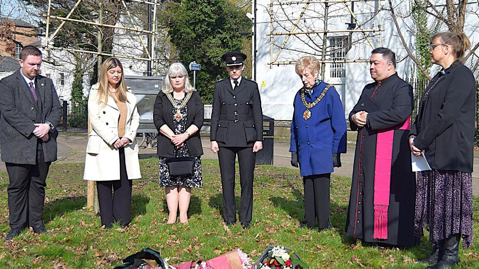 A group of dignitaries, council leaders, police and religious leaders, gathered around some flowers laid on the floor