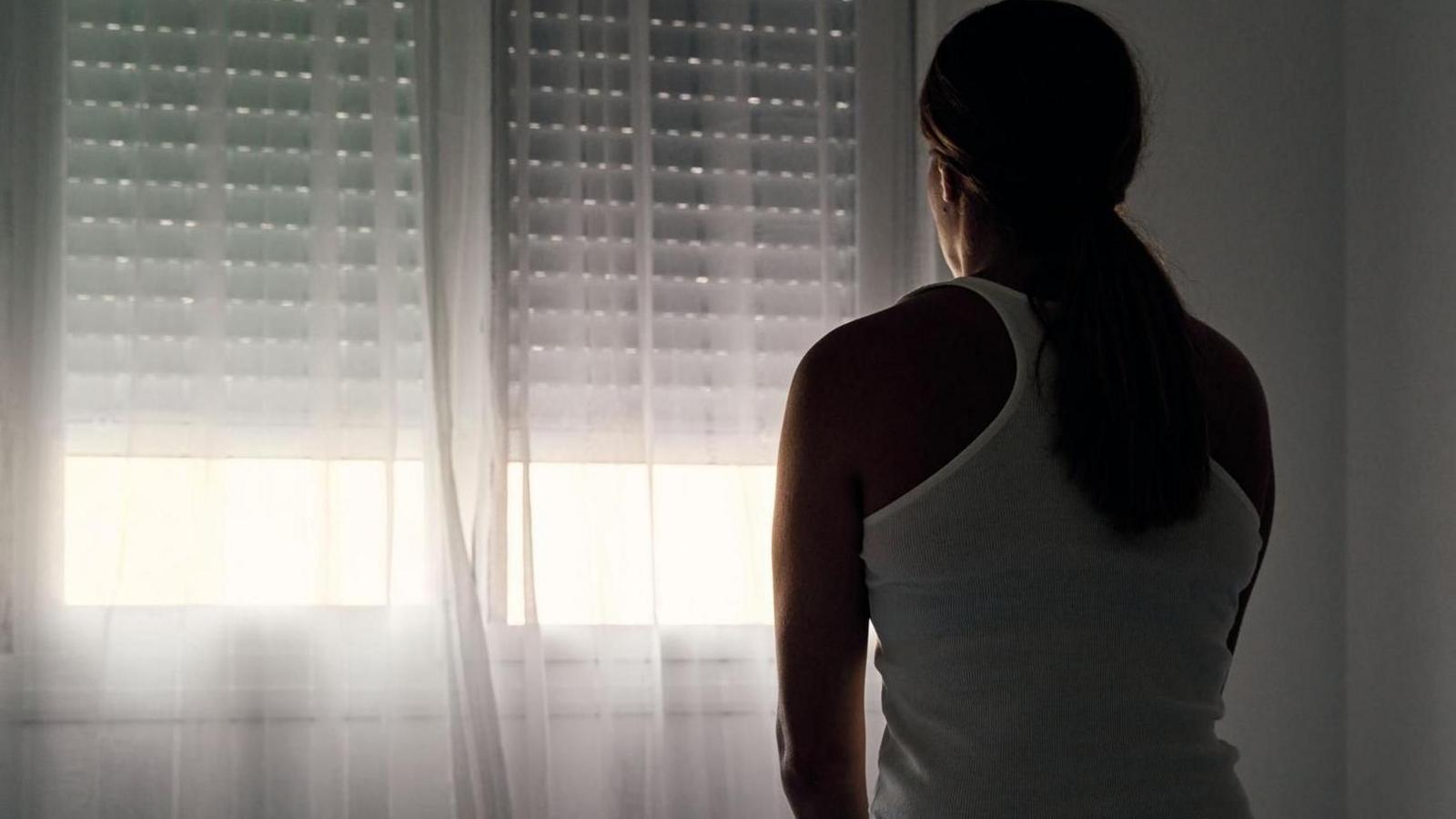 A stock image of a woman sitting on a bed staring out of a window covered by a net curtain.