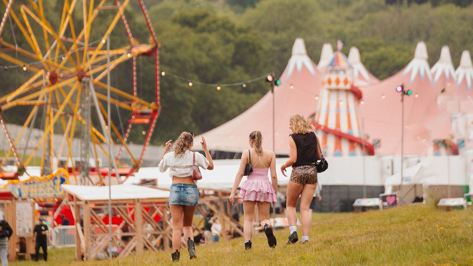 Three women walk towards fairground rides in the distance, including a ferris wheel and helter skelter.