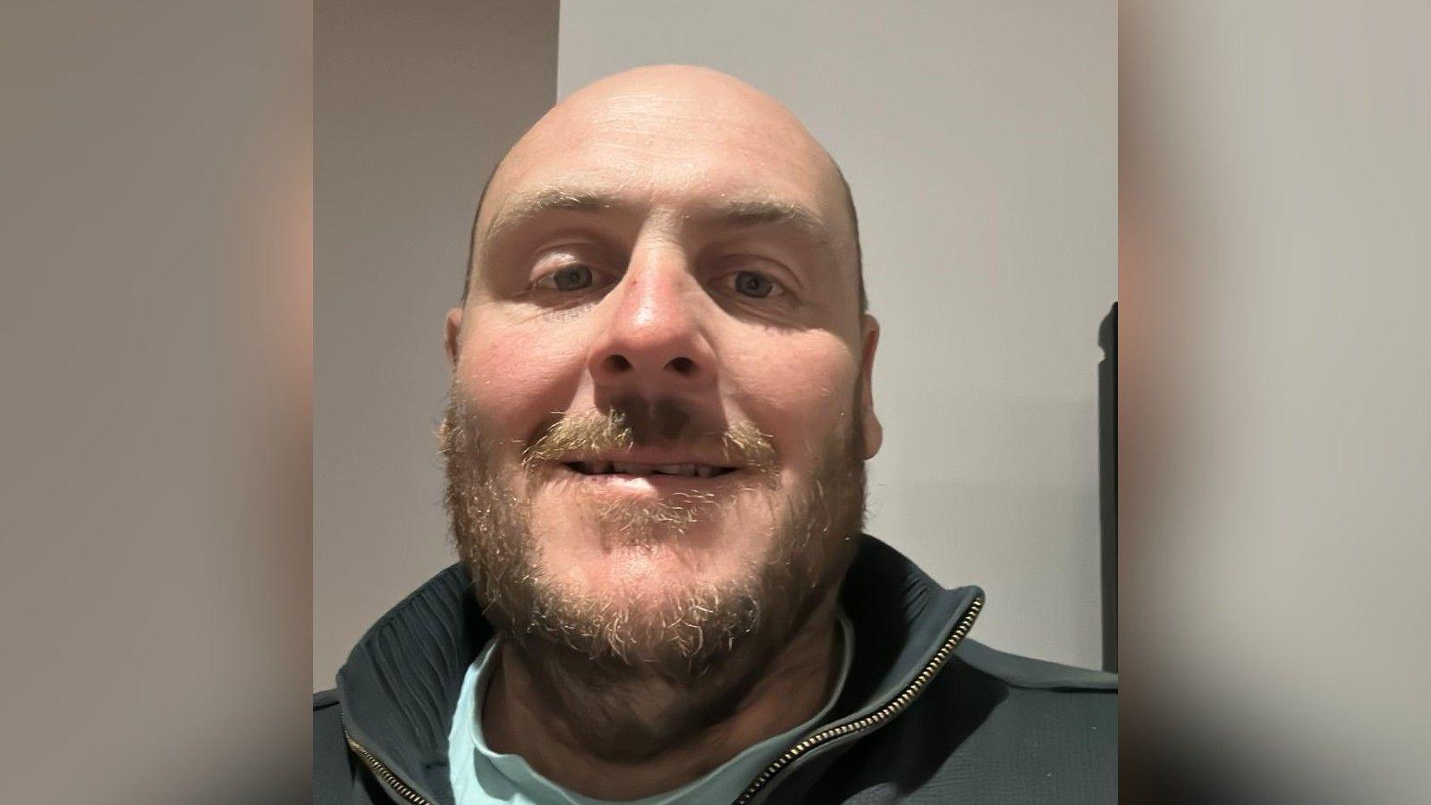 Dave Poulton is pictured smiling in a selfie. He has a dark blond beard and is wearing a high neck collar jumper.