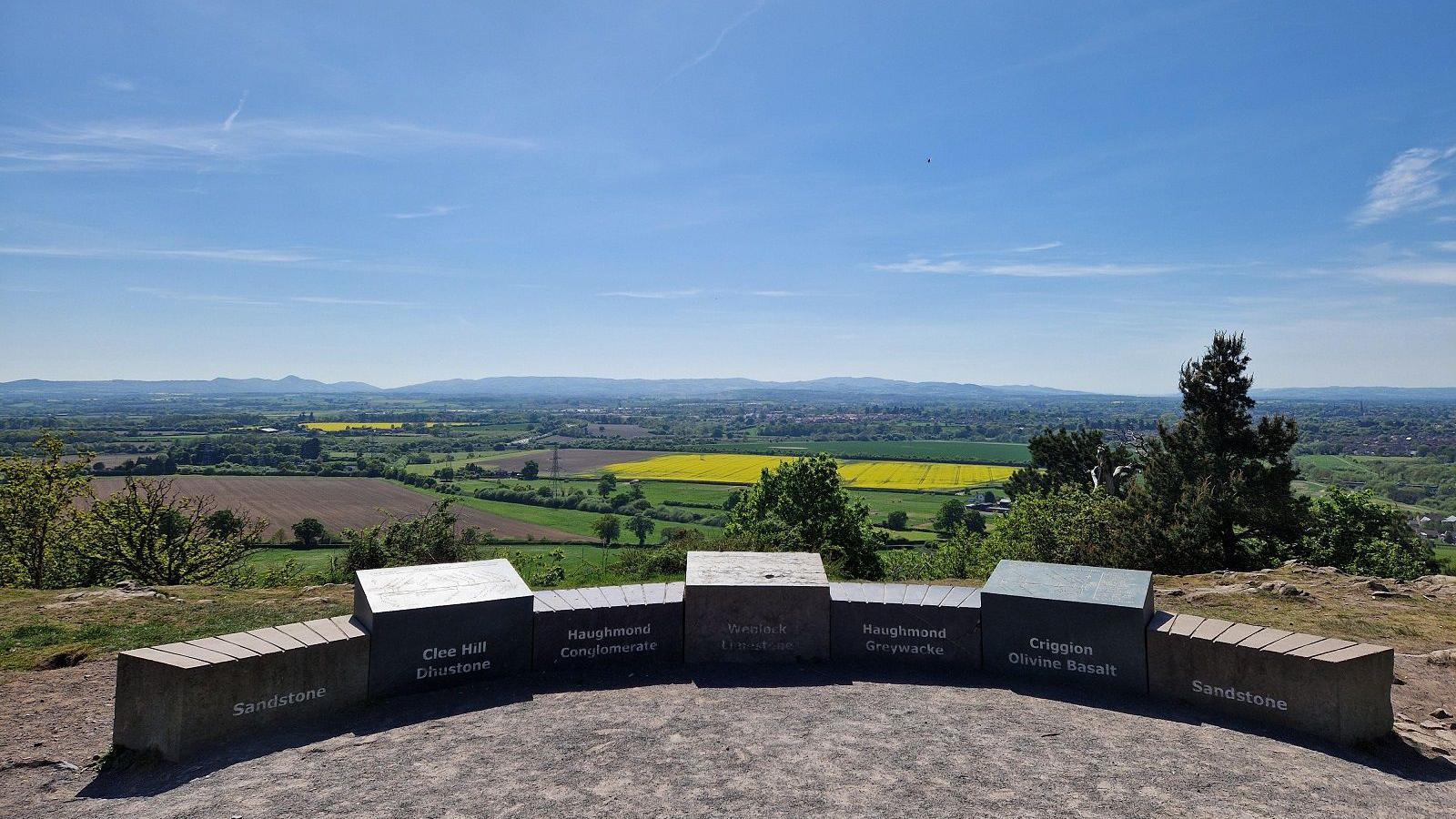 A view from a hilltop showing rolling fields leading away from a hill. In front of them are several blocks used as seating area with words written on them including "Clee Hill" and "Sandstone.