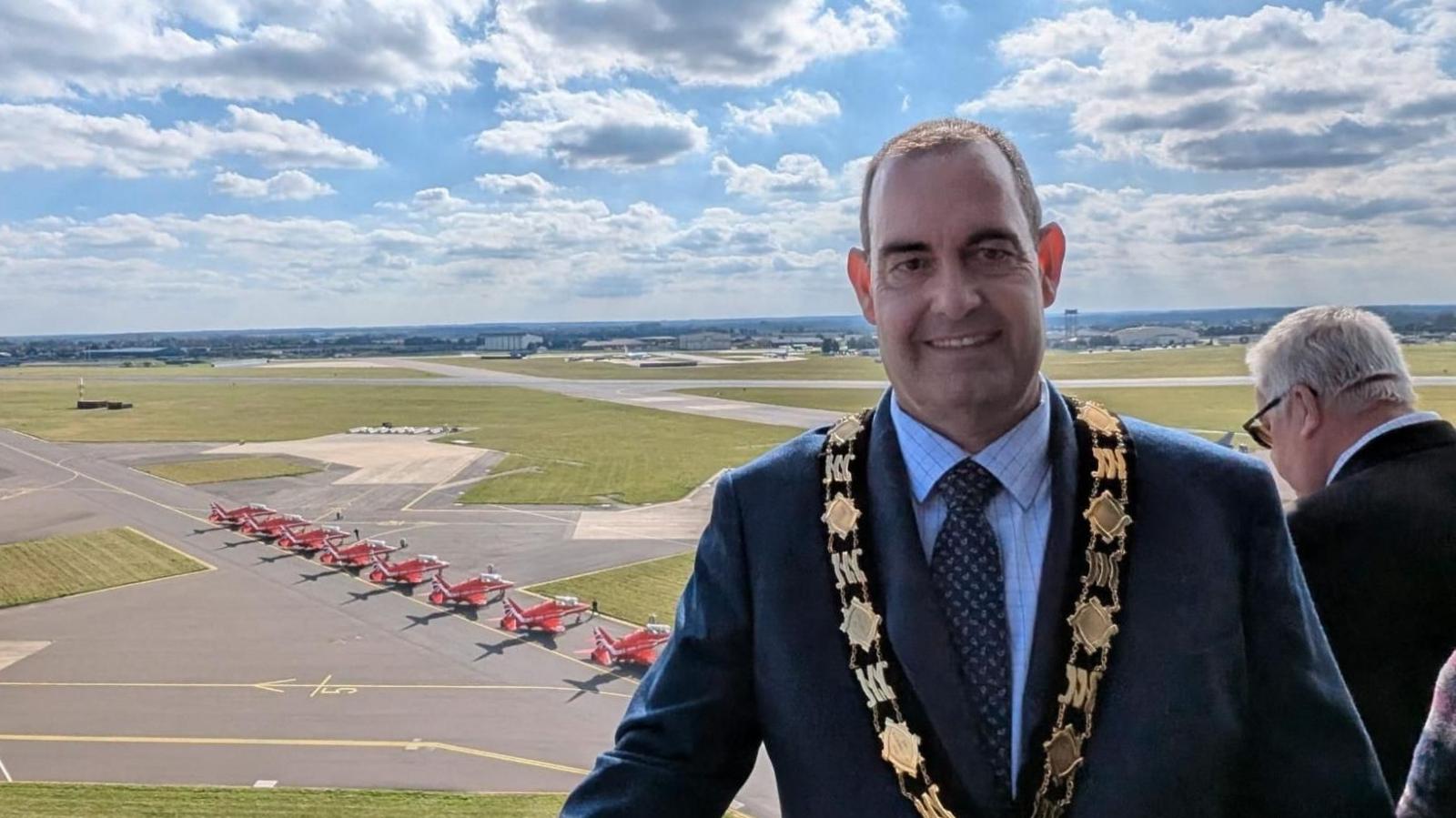 Russell Leaman is standing above a military airfield with eight red aeroplanes on the ground. He is wearing a suit and his mayoral chain. 