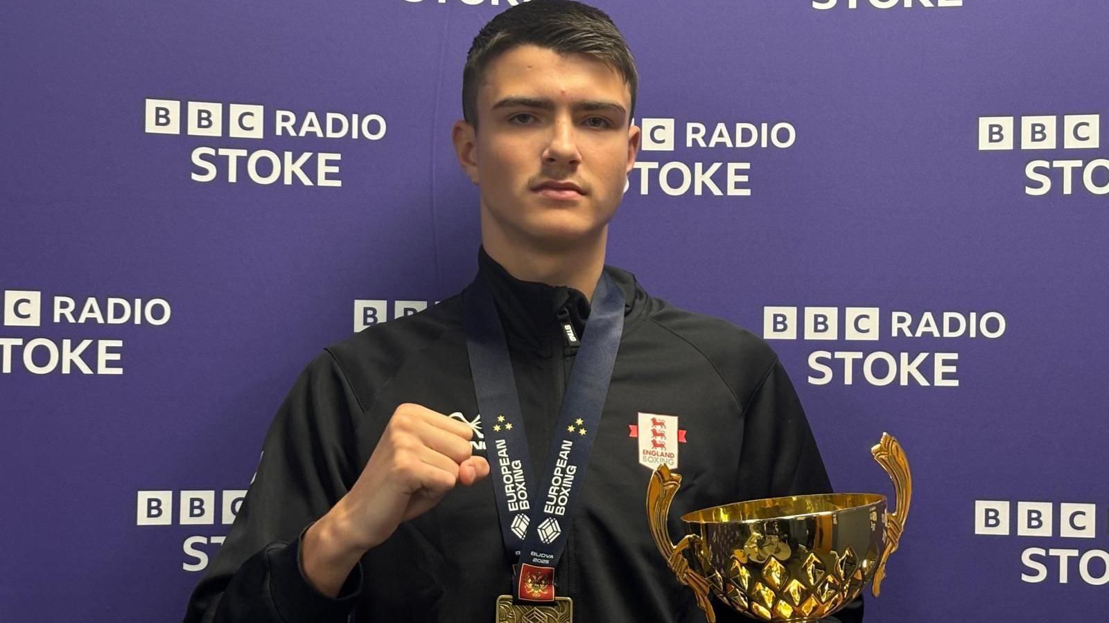 A teenage boy with short black hair, a black top and a medal around his neck is holding his right hand up in a fist while holding a gold trophy in his left hand. He is standing in front of a purple background with "BBC Radio Stoke" in white letters.