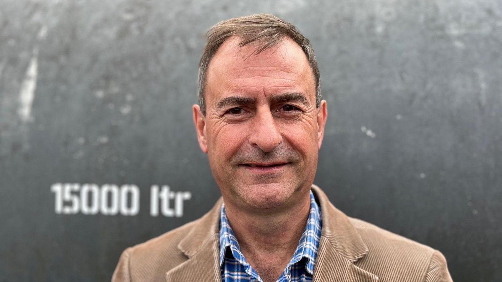 Paul Lewin wearing a brown suit jacket with a blue and white plaid shirt. He has short light brown hair and is clean shaven. He is looking at the camera and is standing in front of a water tank.