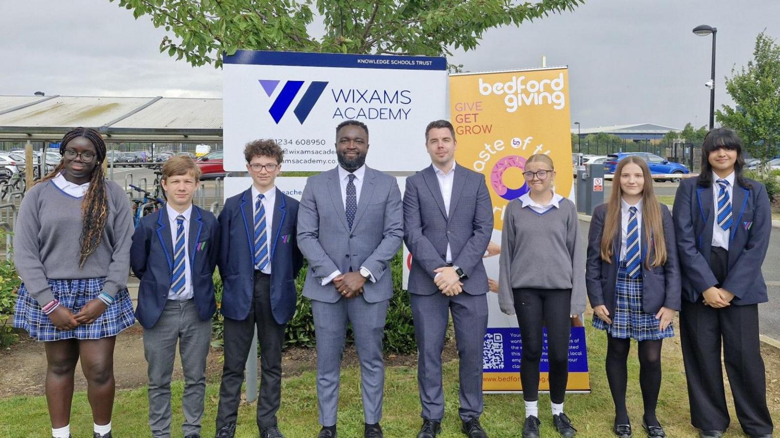 Six pupils standing outside their school, with two adults in the middle. All the pupils are wearing grey and blue school uniform, and the men in the middle, grey suits.