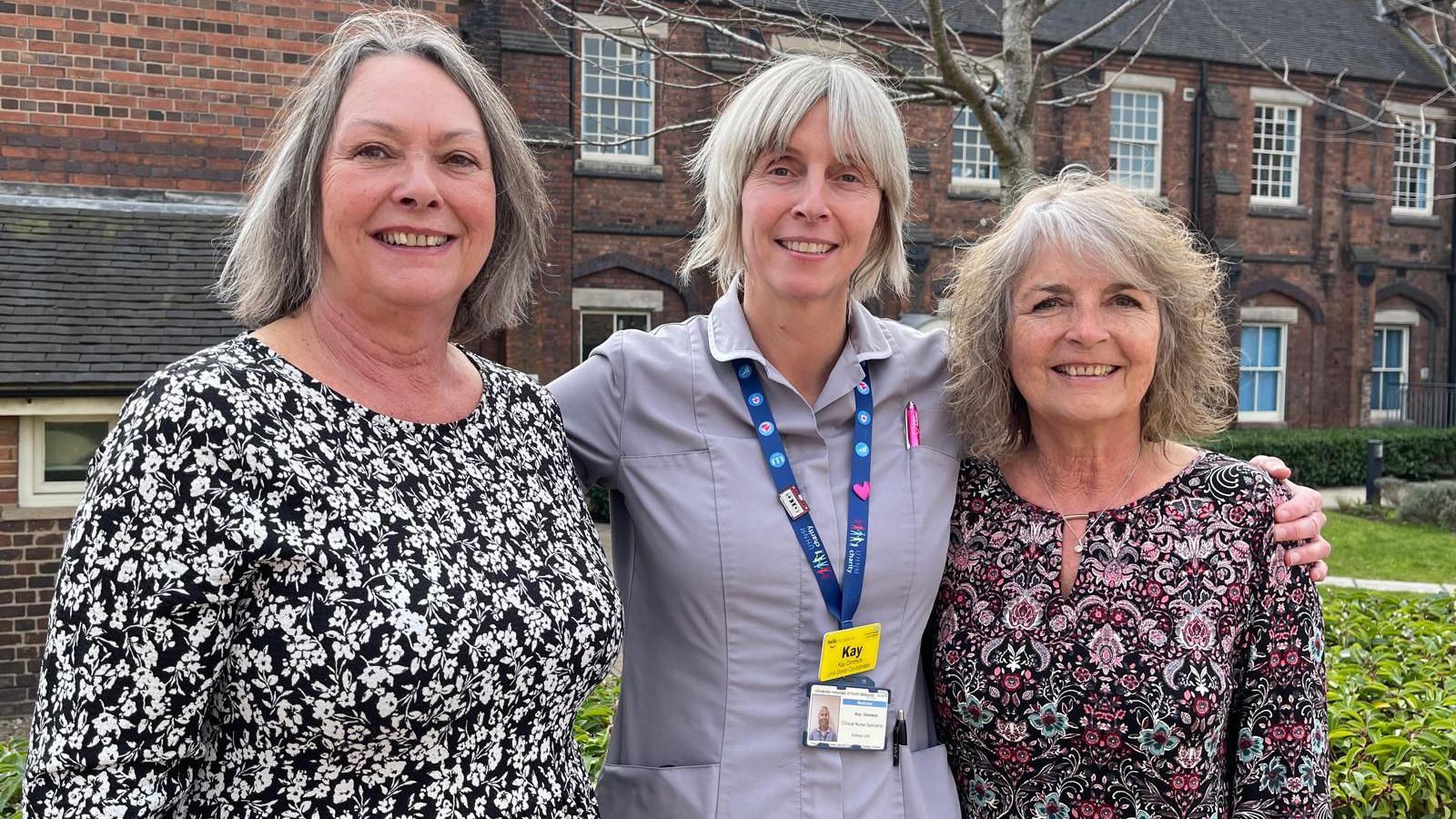 Three women smiling as they stand outside a building. The woman in the middle has her arms around the other two.