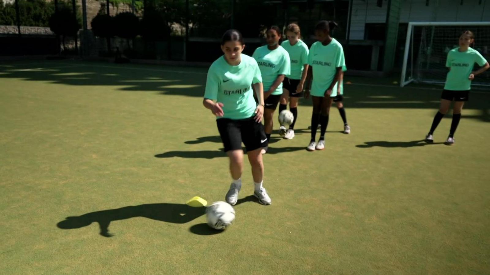 A group of girls in green football jerseys line up to kick a ball at football practice. 