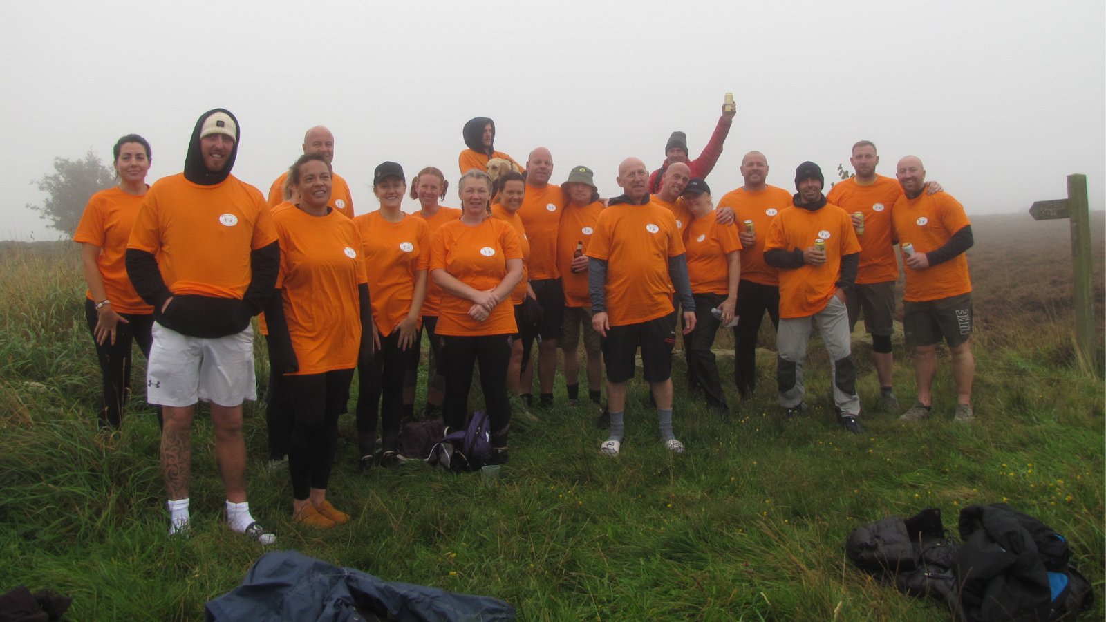 A mixed group of walkers stand on the foggy moorland, wearing bright orange t-shirts over jumpers.