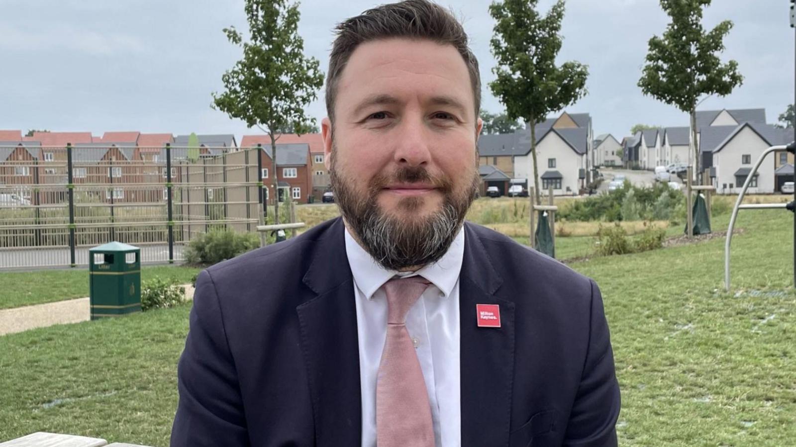 Peter Marland is wearing a navy suit and white shirt with a pink tie. He's standing in a play area near to some new housing, with trees behind him and climbing equipment just visible to the right. Peter has a brown beard and brown hair.