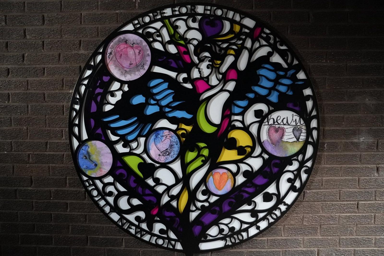 The circular piece of art which shows a dancer with wings with various colourful designs surrounding it. At the top it reads 'Hope for Holly' and at the bottom. The design is largely black and white with bits of pink, green, yellow, blue and purple. It is mounted on a dark, brick wall.