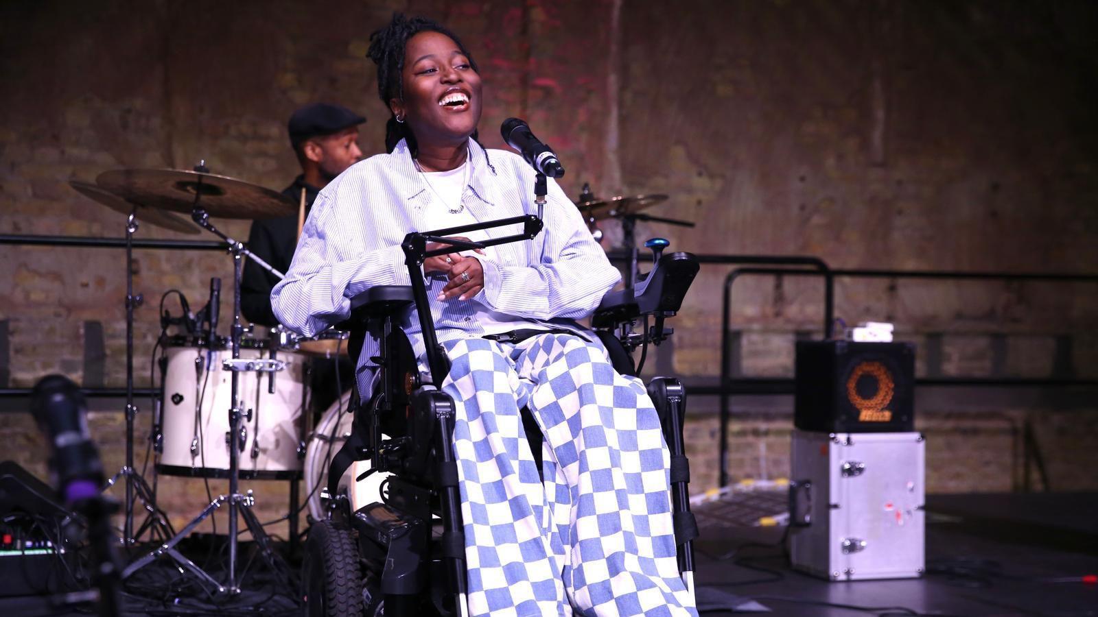 Miss Jacqui, a wheelchair user wearing a lilac shirt and lilac and white checked trousers, performs on stage