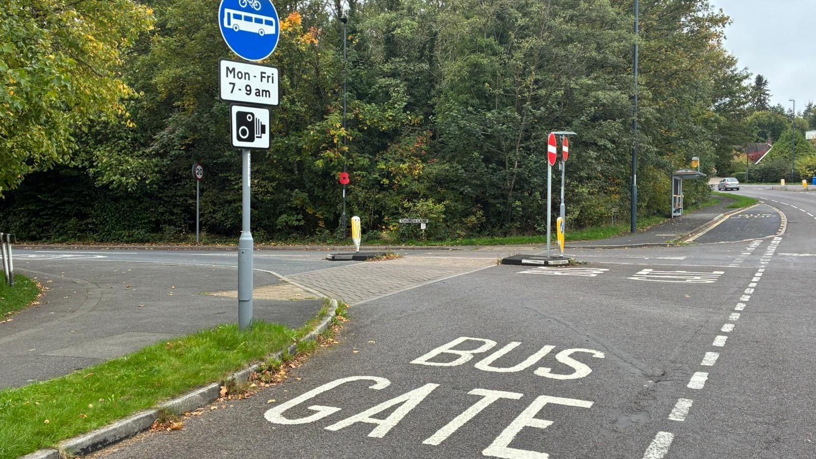The words 'BUS GATE' in white capital letters painted on the road. There is also a blue sign which allows buses and cyclists with a white sign under it which reads 'MON-FRI 7-9 AM.