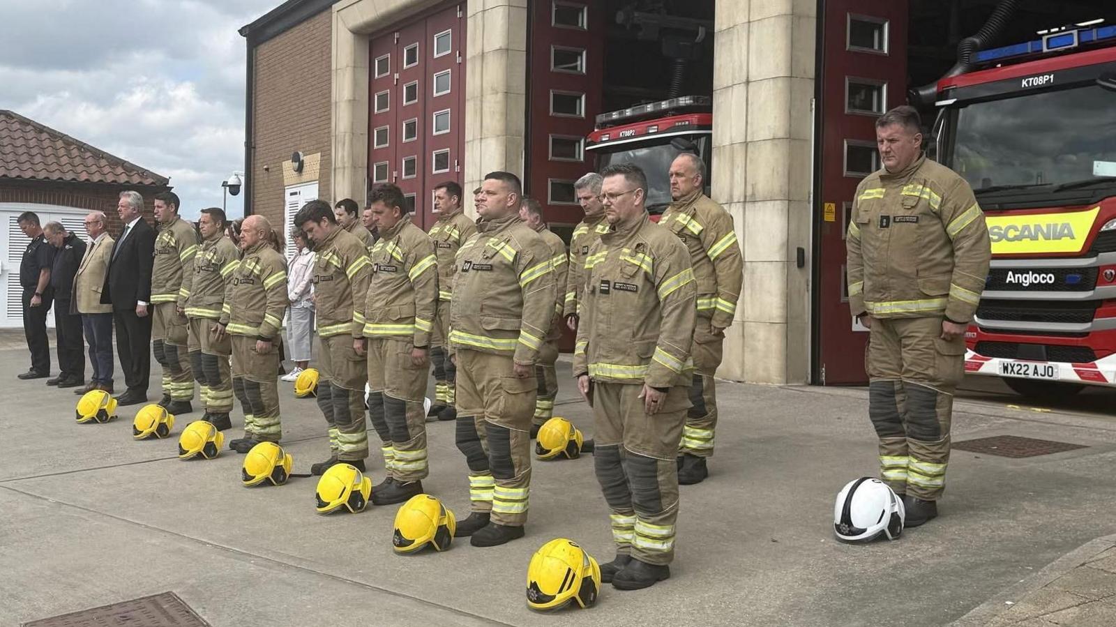 Two rows of firefighters standing outside a fire station. There are about 20 people wearing their firefighting overalls and their yellow helmets are on the floor at their feet.. Behind them the doors are open and two fire engines can be seen.
