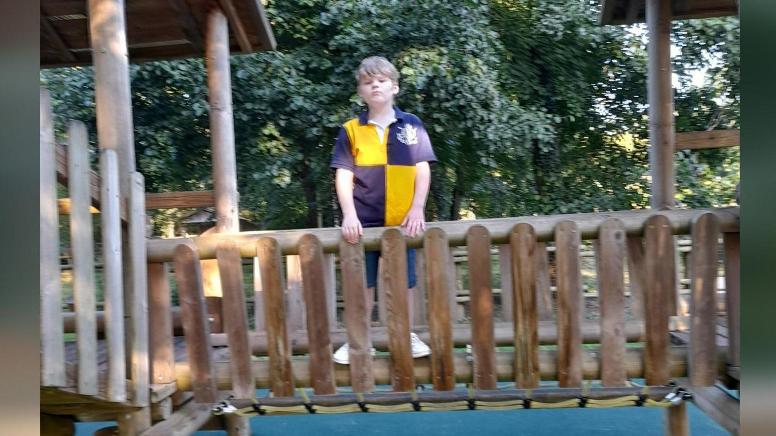 Alex posing for a photograph on the wooden bridge on a climbing frame in a playpark. He is wearing a yellow and blue quartered rugby style top and is looking at the camera. He is not smiling.