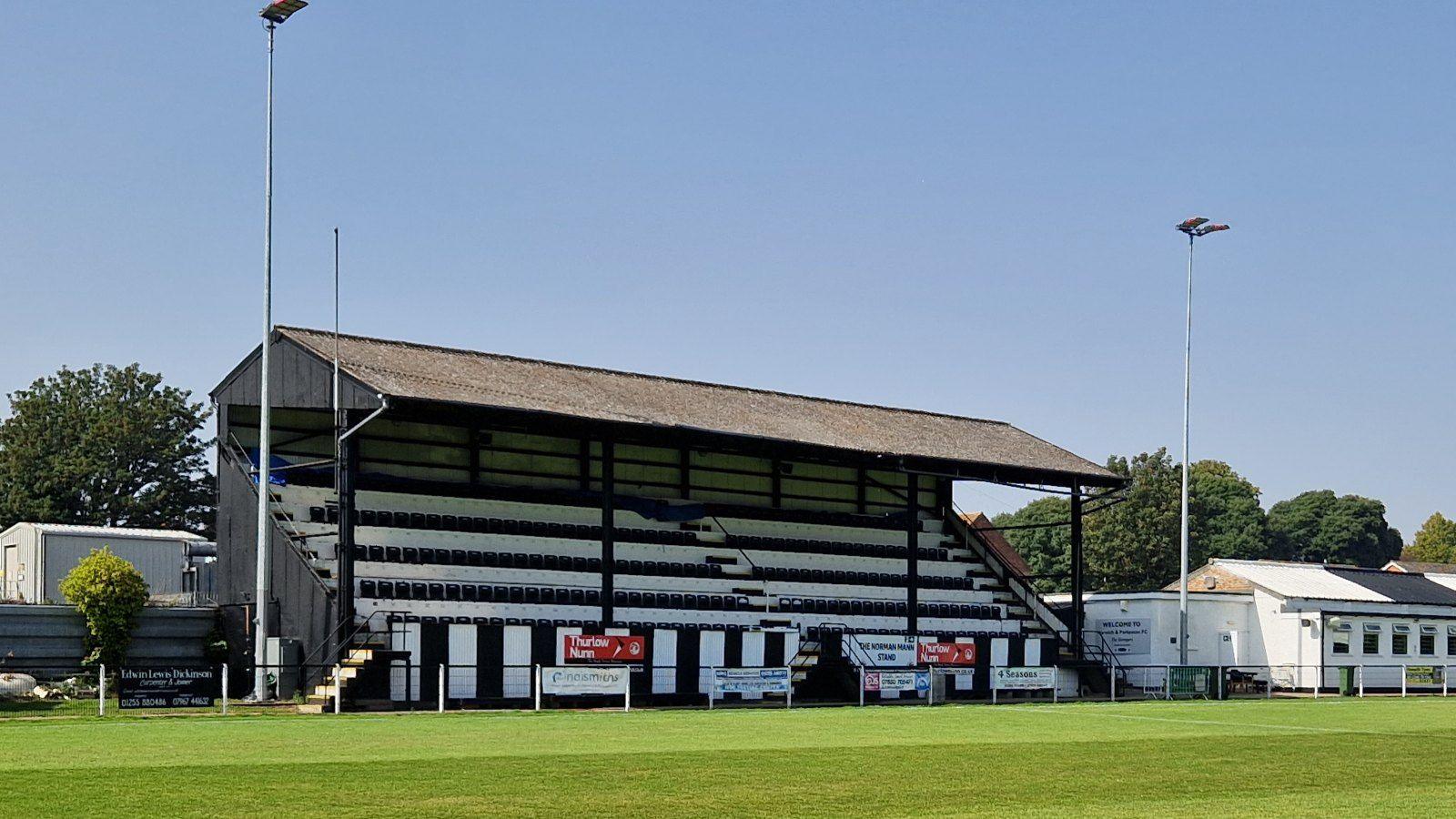 Harwich & Parkeston FC's home ground Royal Oak. It is a steep stand, with black and white seats. The pitch is visible in the foreground. There is a blue sky.