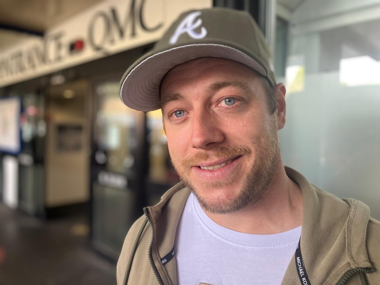 Zak Logalbo pictured outside the QMC hospital entrance. He is wearing a khaki green jumper and white t-shirt, with a green and white cap.
