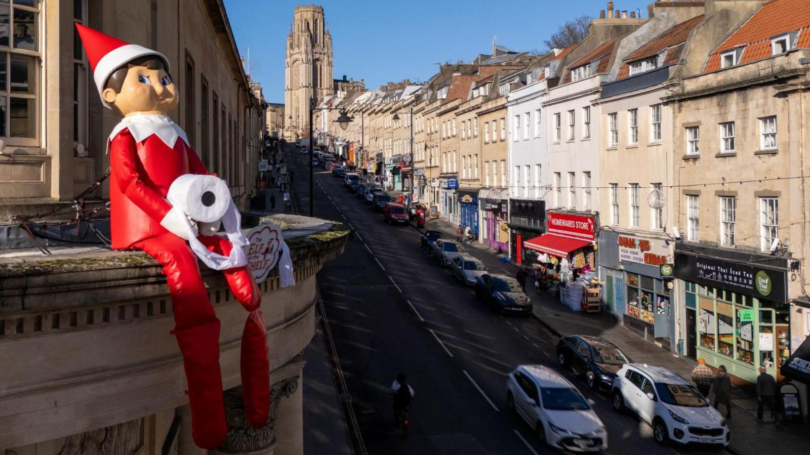 A large Christmas elf in a red and white outfit is pictured on the parapet of a building on the side of Park Street in Bristol. Behind the statue of the elf, the street is visible with traffic on it and cars parked either side. In the distance the Wills Memorial Building can be seen and it is a sunny day with clear blue skies