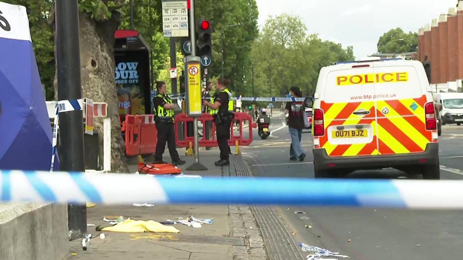 Two officers talk to each other behind a cordon, with a red paramedic kit and medical wrappers lying on the ground in front of them