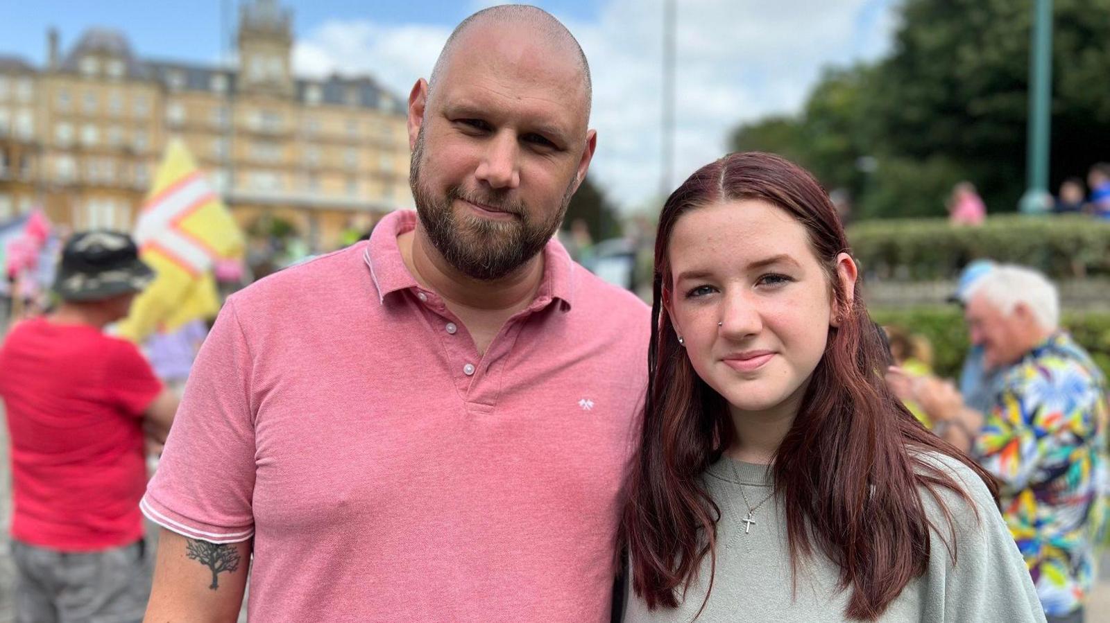 A bald man with a beard wearing a pink polo neck and a young teenage girl with dark red hair wearing a grey sweatshirt smiling at the camera. The town hall building and a couple of other protesters are in soft focus behind them.