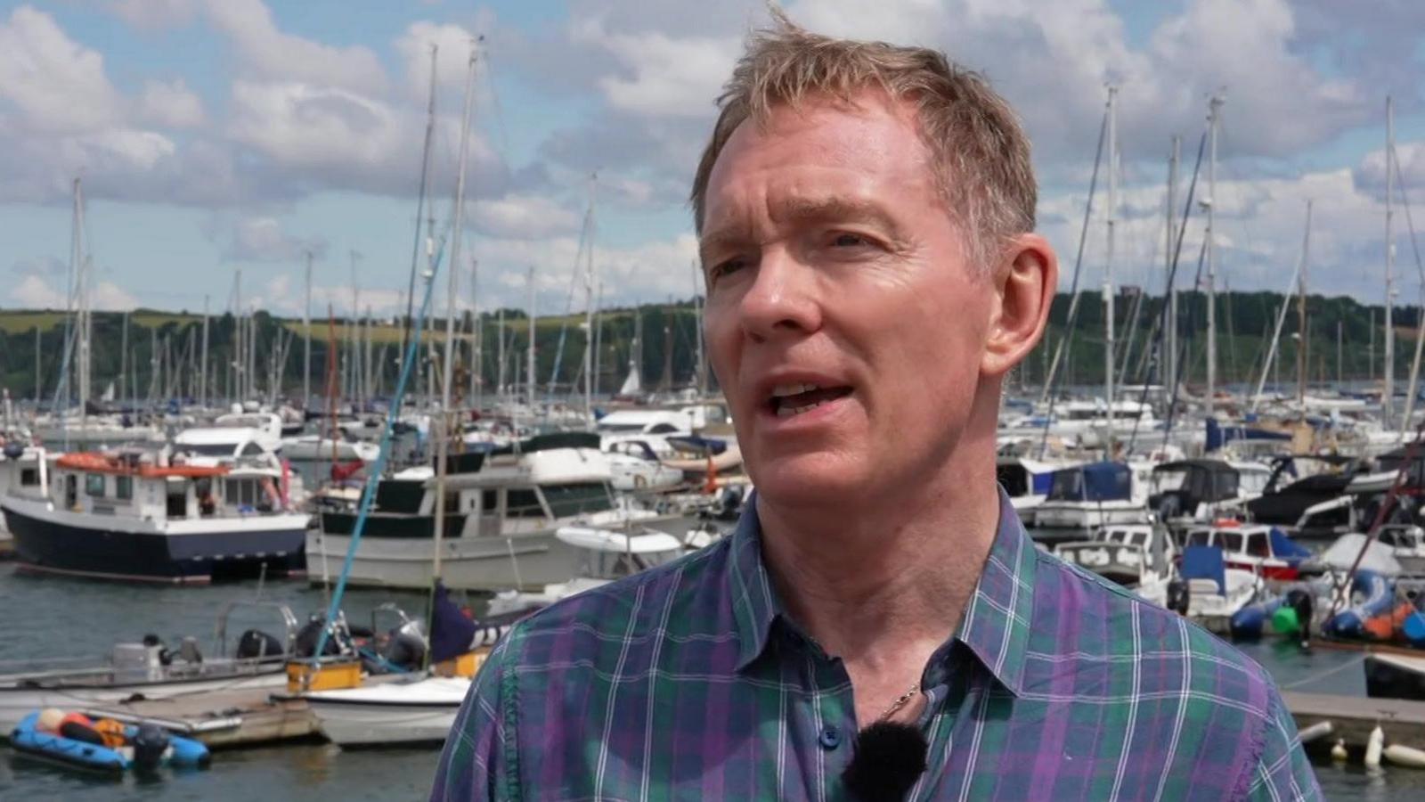Tourism Minister Sir Chris Bryant wears a green and purple tartan style shirt while stood in a marina in Mylor, Cornwall. There are dozens of boats and yachts behind him.