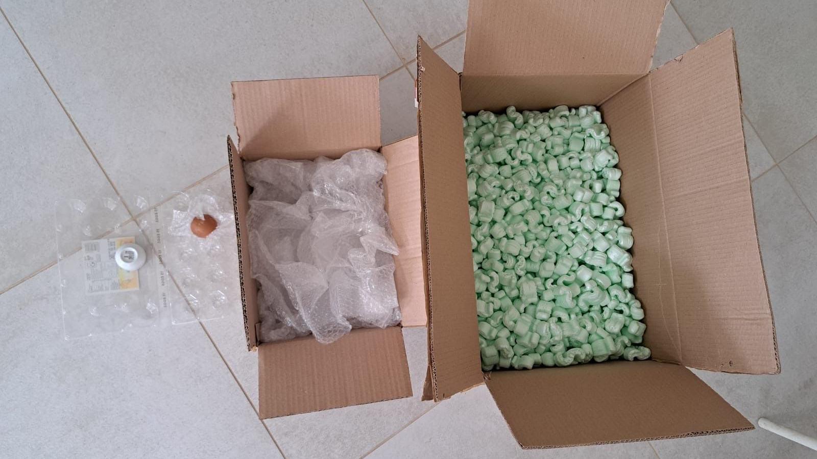 An egg in a clear plastic supermarket egg tray sits on a tiled floor. Next to it is a cardboard box filled with bubble wrap. Next to that is a larger cardboard box filled with green polystyrene packing peanuts.