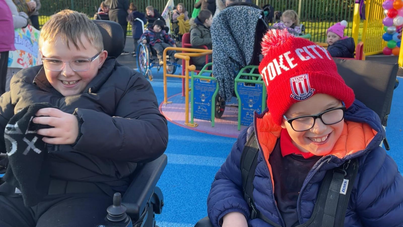 Two boys in wheelchairs sit in a colourful playground. Behind them children are playing on a playground roundabout. The boy on the left has blonde hair and wears glasses and a black puffer coat. He is holding a black hat in his hands. The boy on the right also wears glasses but wears a red and white Stoke City hat and a blue puffer coat with orange lining. The zip is undone showing his navy school jumper and red polo shirt. They are both smiling.