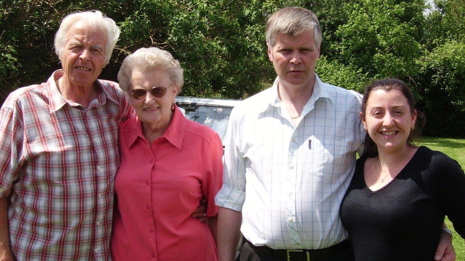 A family photo of two grandparents, an uncle and granddaughter standing in a row. The elderly man is wearing a checked shirt, next to his wife who is wearing a pink shirt and sunglasses. In the middle is a man with grey hair and a white shirt, on the right is a woman with dark hair wearing a black top who is smiling. They are standing in a garden on a sunny day.