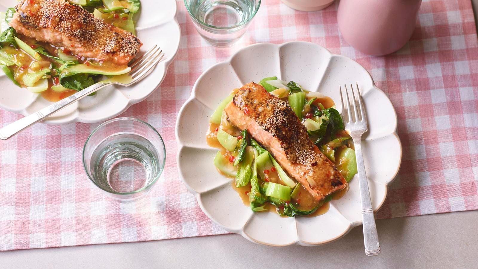 Two white scalloped bowls shot from above. Each contains a bed of ribbon veg in a red sauce with a fillet of salmon encrusted with seeds on top. The table cloth is pink gingham and there are two glasses of water.