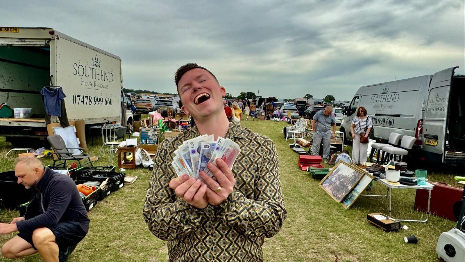 A man standing in the middle of a car boot sale with a wad of cash notes in his hand, his head is tipped back with a look of bliss on his face.