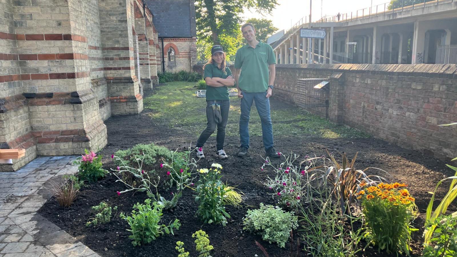 Woman wearing green T-shirt, cap and dark jeans with man wearing green T-shirt with blue jeans stand on a patch of soil behind plants, next to a paved area. There is a brick and stone church building to one side of them and a brick wall and another building to the other.