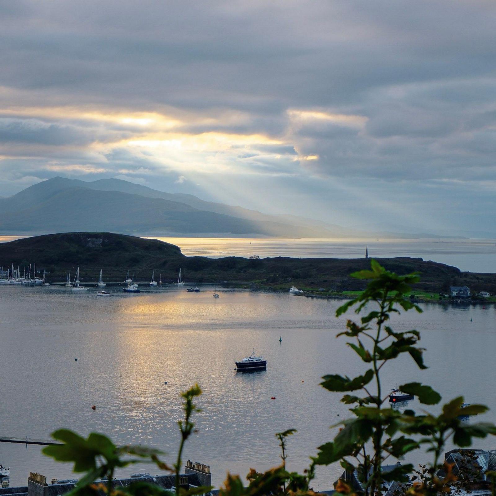 Light shines from between clouds over a large expanse of water with tiny boats on the far side.