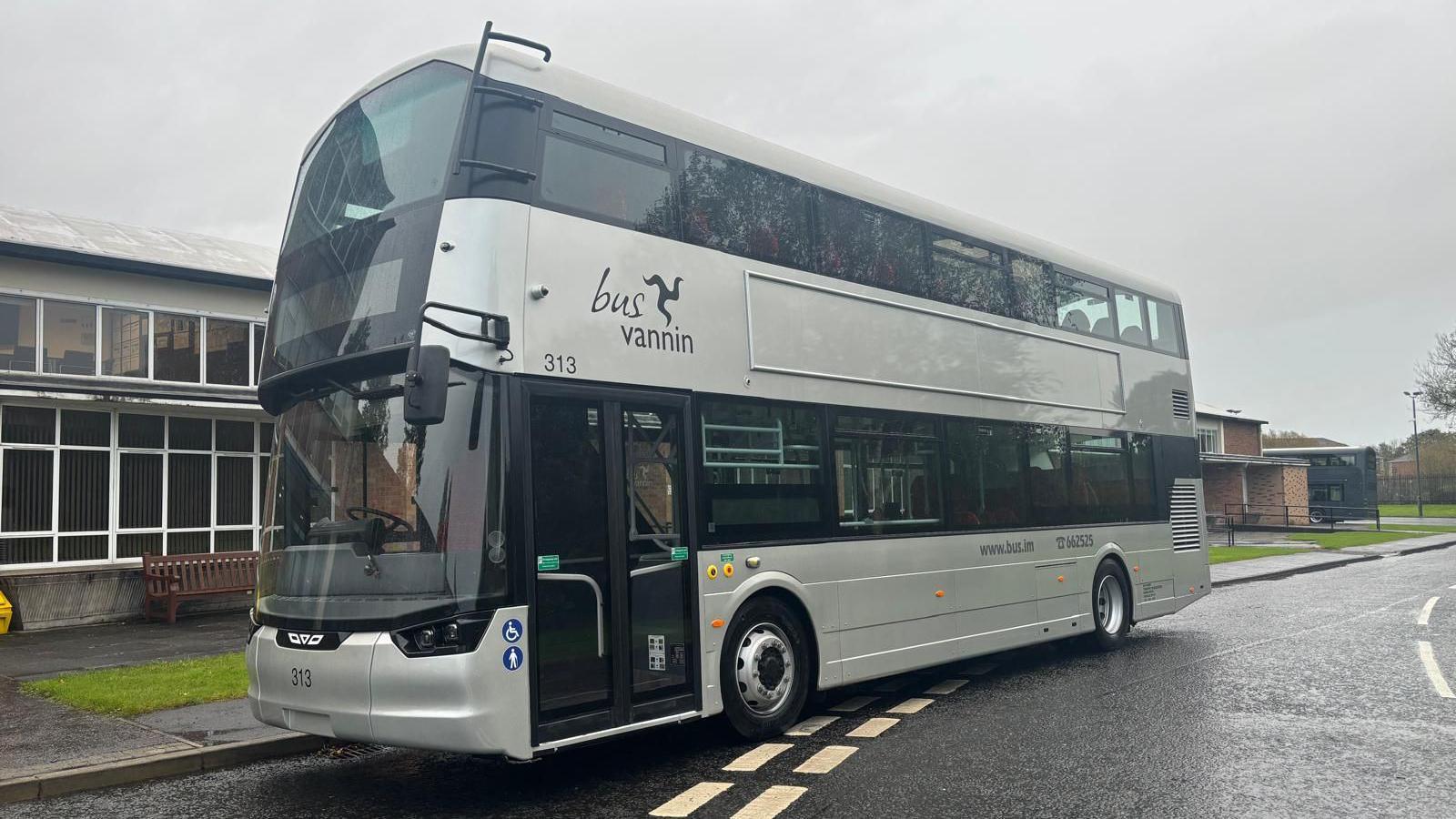 A silver double-decker bus with Bus Vannin above the vehicle's double entrance doors. It is parked next to a long building with lots of white-framed windows.