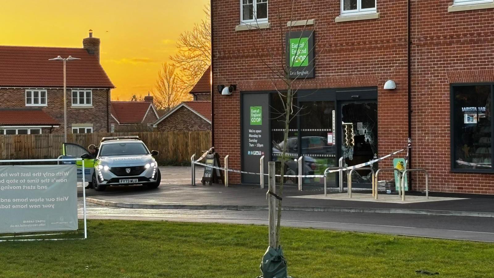 A wider view of the police cordon outside the Co-op supermarket. A police officer steps out from a police car that is parked up outside. The sun is rising behind some houses to the right of the image.