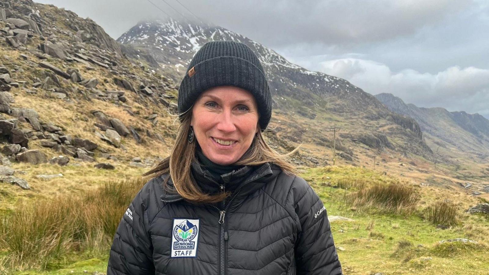 Llun o Angela Jones, Pennaeth Partneriaethau Awdurdod Parc Cenedlaethol Eryri yn edrych ar y camera. Mae hi wedi gwisgo'n gynnes mewn cot a het ac yn sefyllar y mynyddoedd.