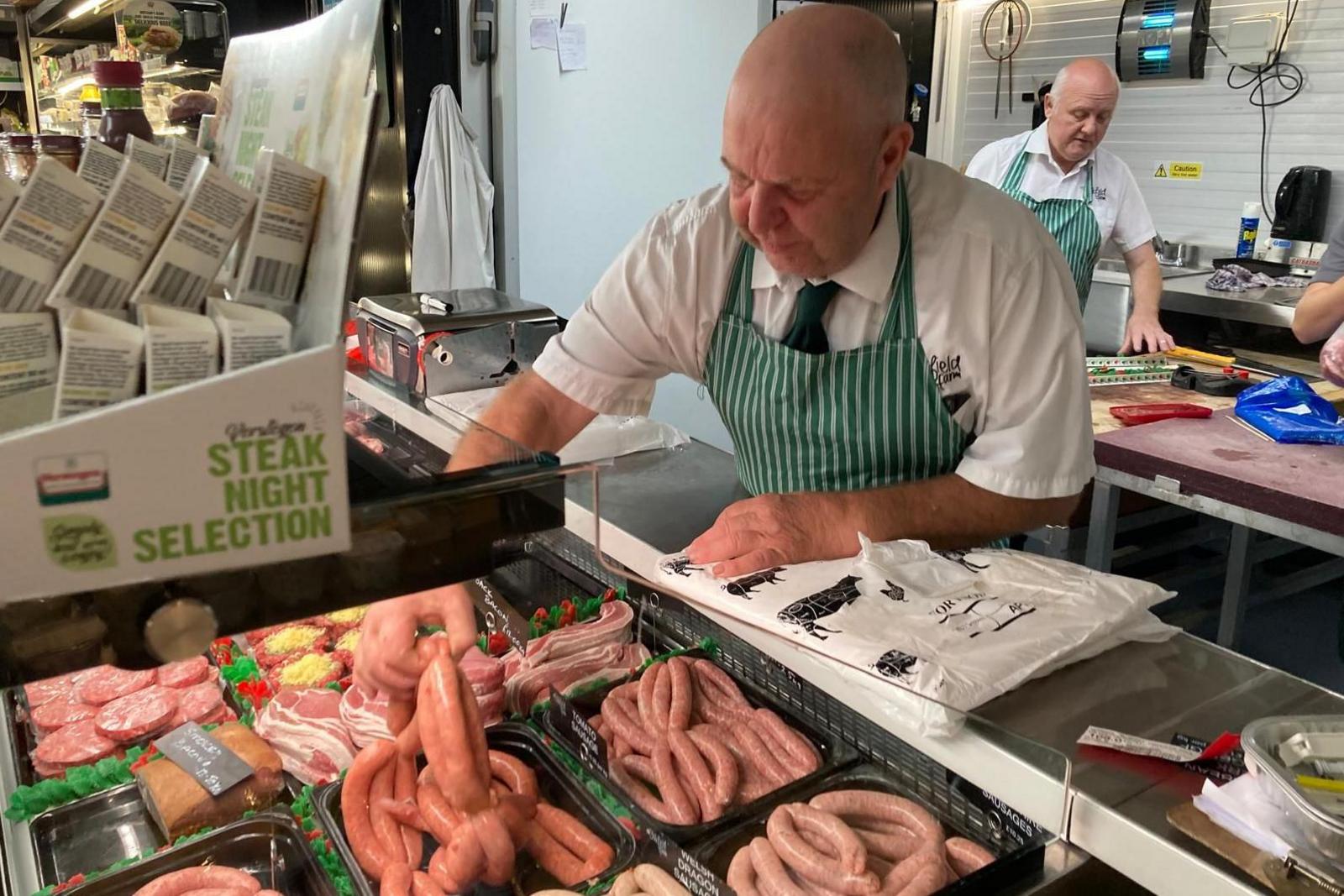 A bald man wearing a white shirt, dark green tie and green apron is in leaning over a counter in a butchery and is picking up sausages. There are other meats on the counter and there is one other worker in the background working on a chopping counter.
