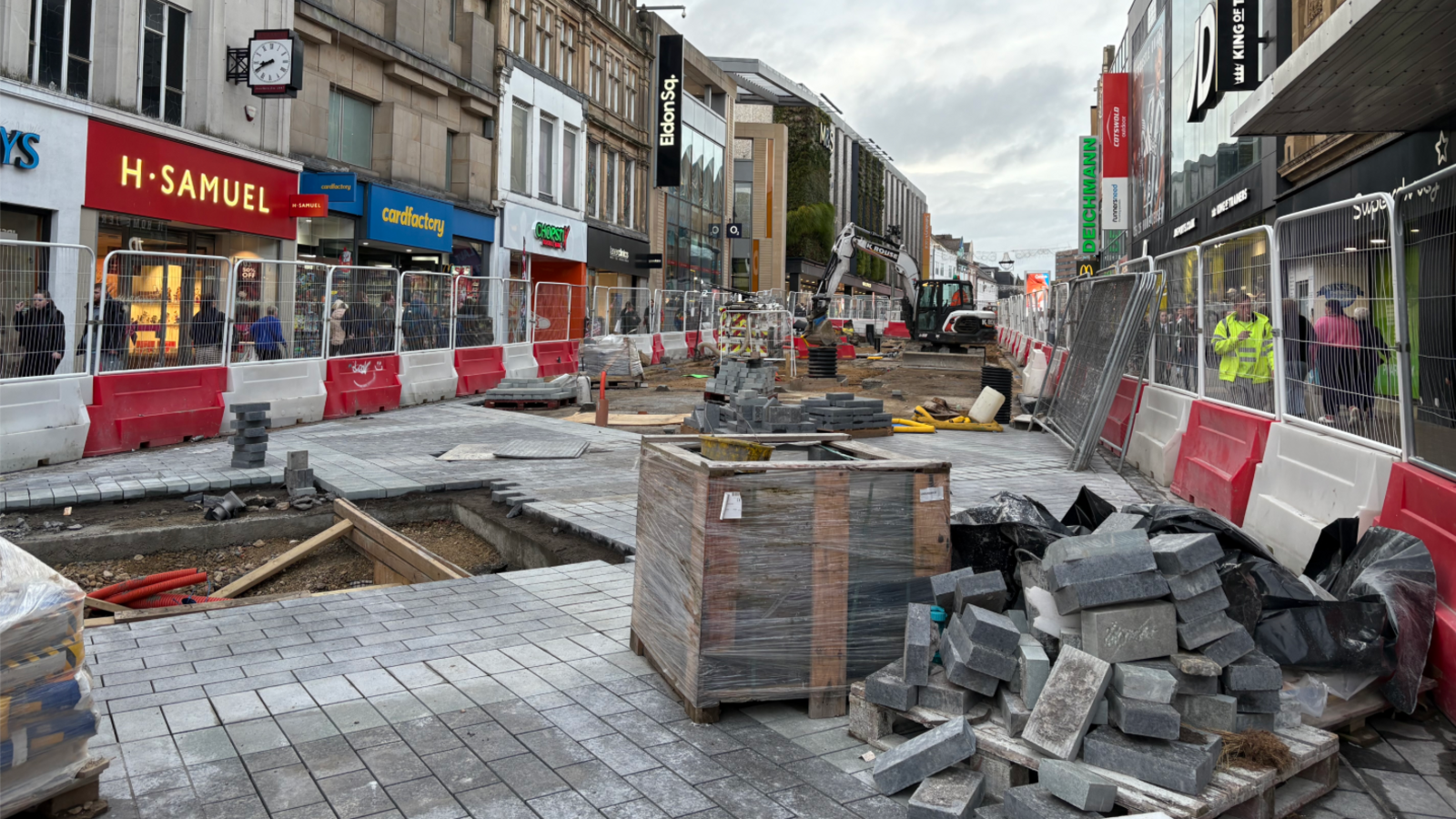 The majority of Northumberland Street is taken up with construction work. There shops are visible on either side, with railed fencing blocking off most of the pathway. Within this fence, sections of the paved floor have been removed, with piles of bricks stacked up, and a digger is visible in the background.