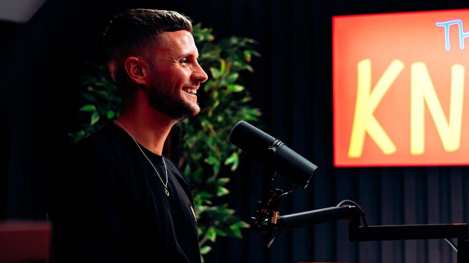 A man with short dark hair, a black shirt and a necklace smiles as he sits in front of a microphone in a studio. Behind him is an orange sign and a large plant.
